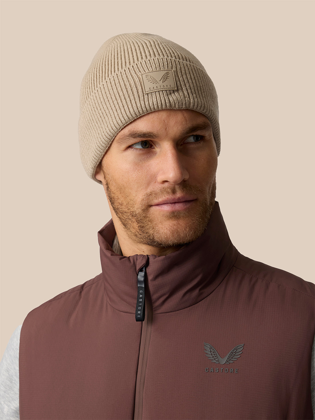 A man wearing a beige ribbed knit beanie with a winged logo patch and a brown zip-up vest with a matching winged logo on the chest, set against a solid beige background.