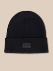 A black ribbed knit beanie hat with a folded cuff and a small dark gray winged logo patch on the front, set against a solid beige background.