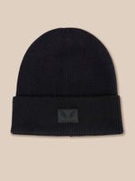 A black ribbed knit beanie hat with a folded cuff and a small dark gray winged logo patch on the front, set against a solid beige background.