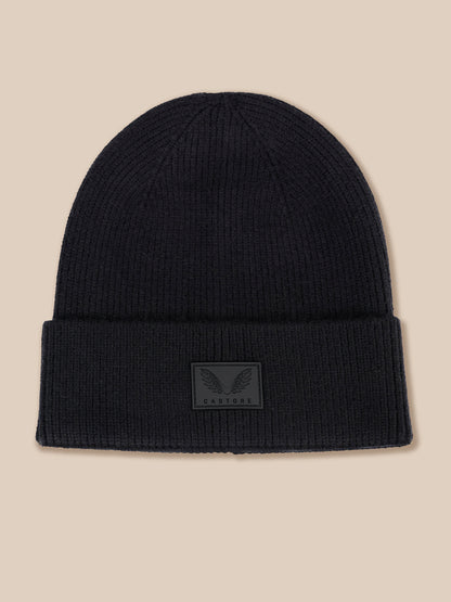 A black ribbed knit beanie hat with a folded cuff and a small dark gray winged logo patch on the front, set against a solid beige background.