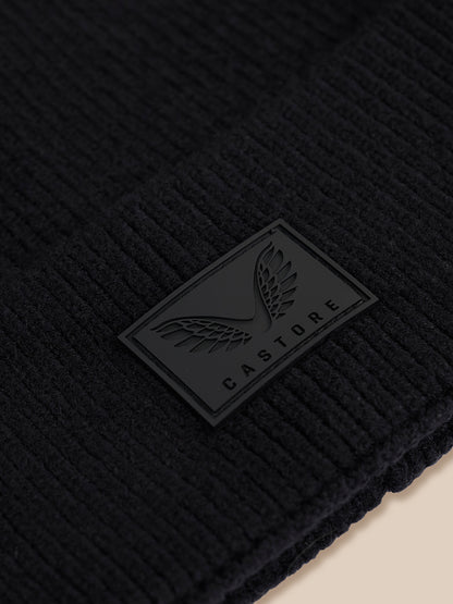 Close-up of a black ribbed knit beanie with a black rubber patch featuring a winged logo and the brand name CASTORE.