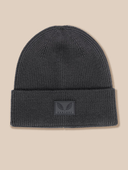 A dark gray ribbed knit beanie hat with a folded cuff, featuring a small rectangular embossed patch on the front with a winged logo and the brand name CASTORE.