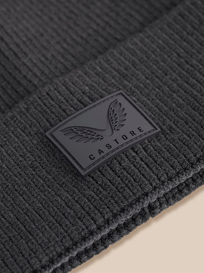 Close-up of a dark charcoal gray ribbed knit beanie with a black rectangular Castore logo patch on the cuff.