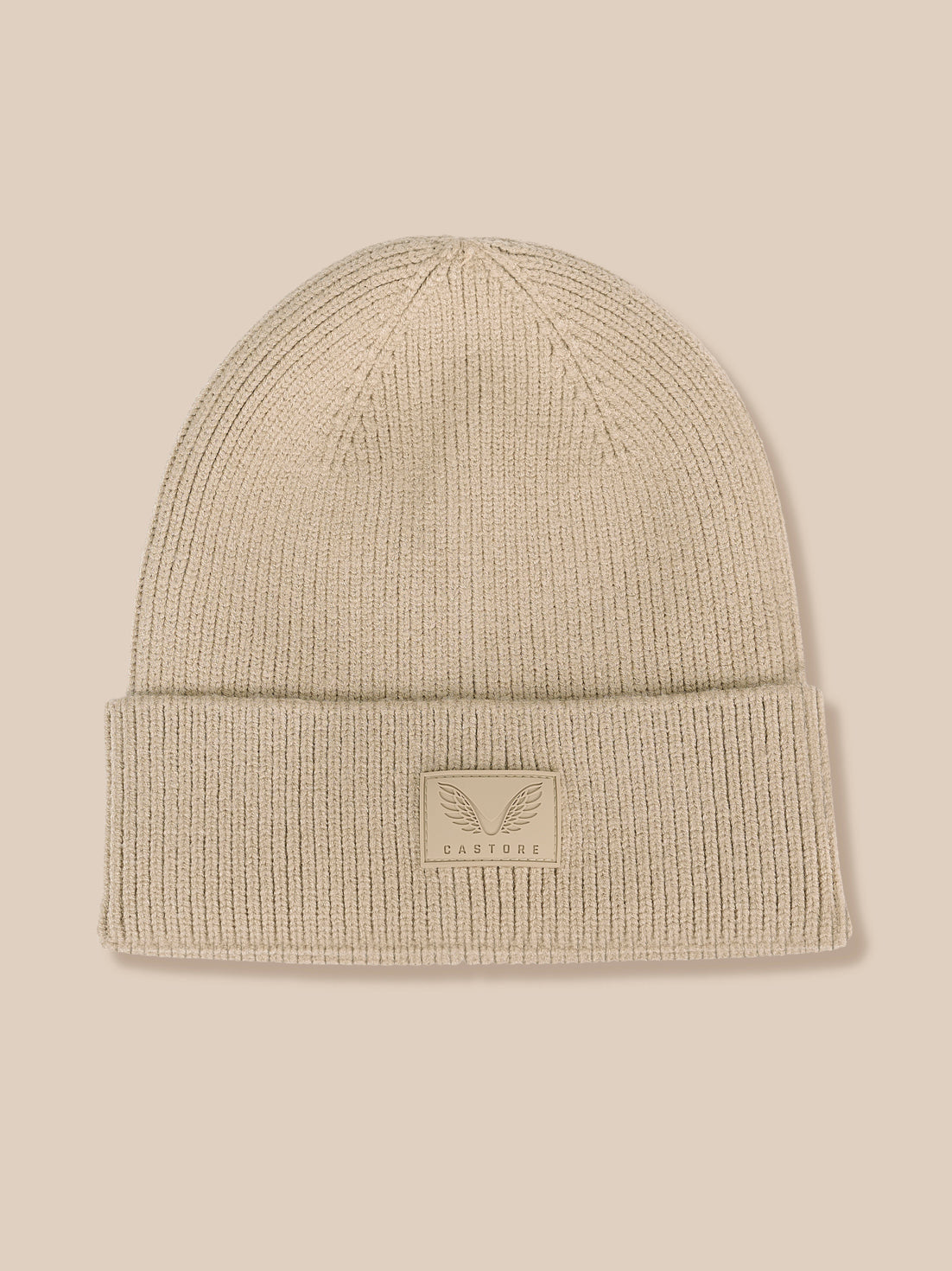 A light beige ribbed knit beanie hat with a folded cuff and a small rectangular logo patch on the front, shown against a solid beige background.