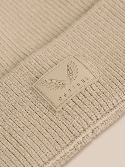 Close-up of a ribbed knit beanie in a warm beige or cream tone, featuring a rectangular leather patch with a winged logo and the word CASTORE.