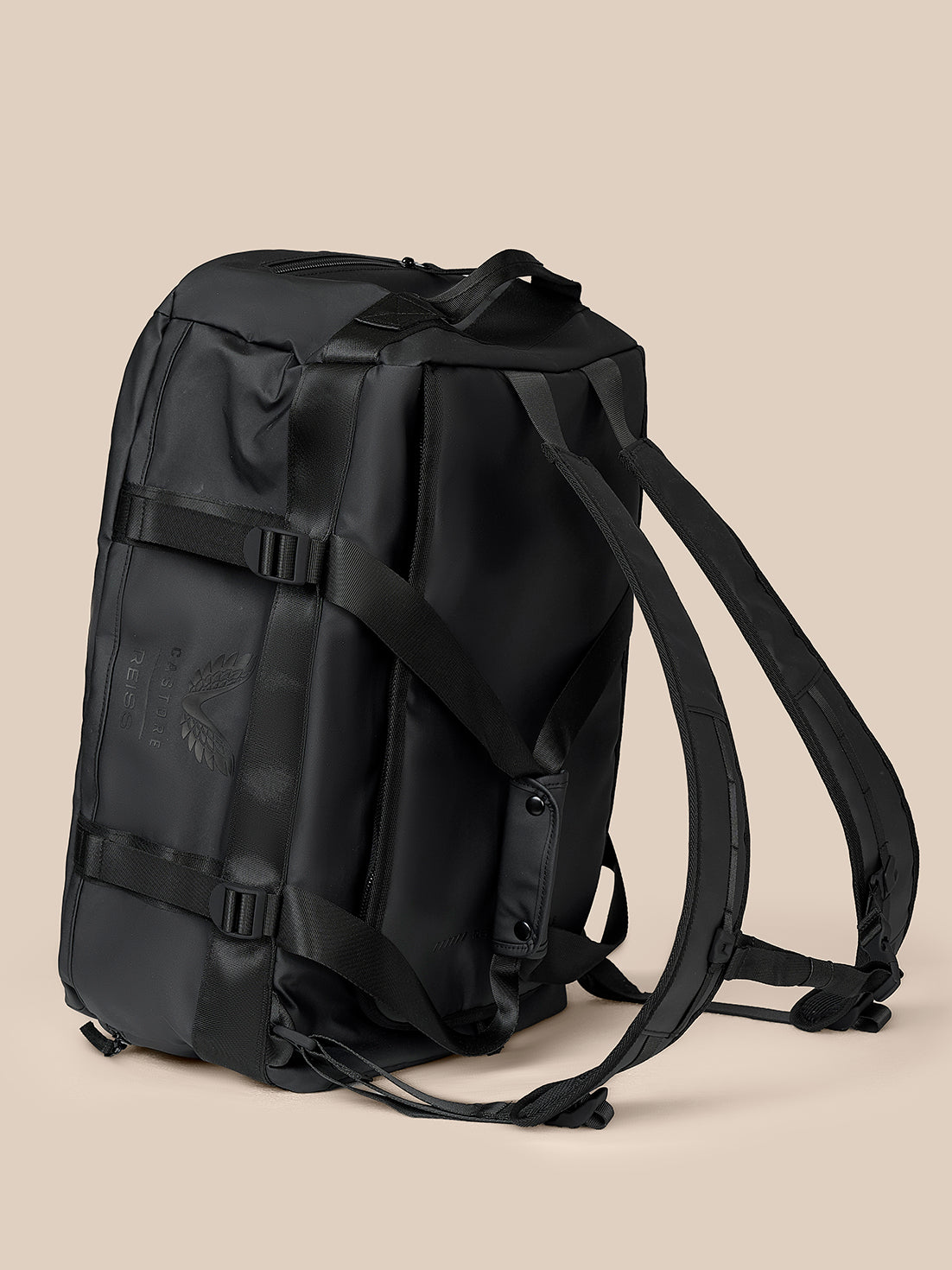 A black Castore Reiss backpack with a tonal dark gray wing logo and brand name printed on the front, set against a solid beige background.