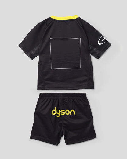 A toddler black short-sleeve jersey and matching shorts set from Dyson, shown flat-laid on a light gray background. The jersey features a bright yellow collar, a white outlined square on the back, and a white circular logo on the sleeve.
