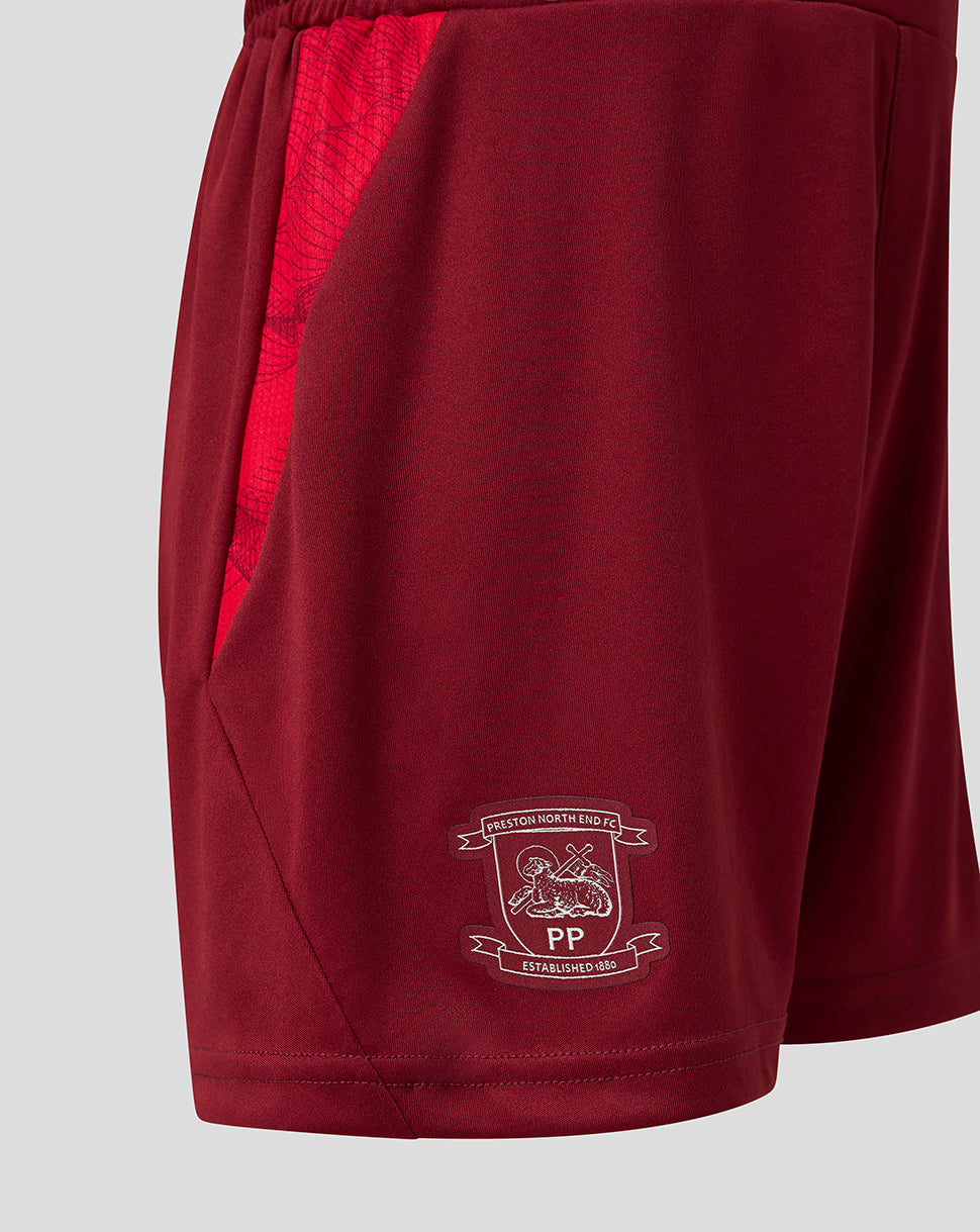 Close-up of maroon athletic shorts with a red patterned side panel and a white printed football club crest on the lower left leg, shown against a light gray background.