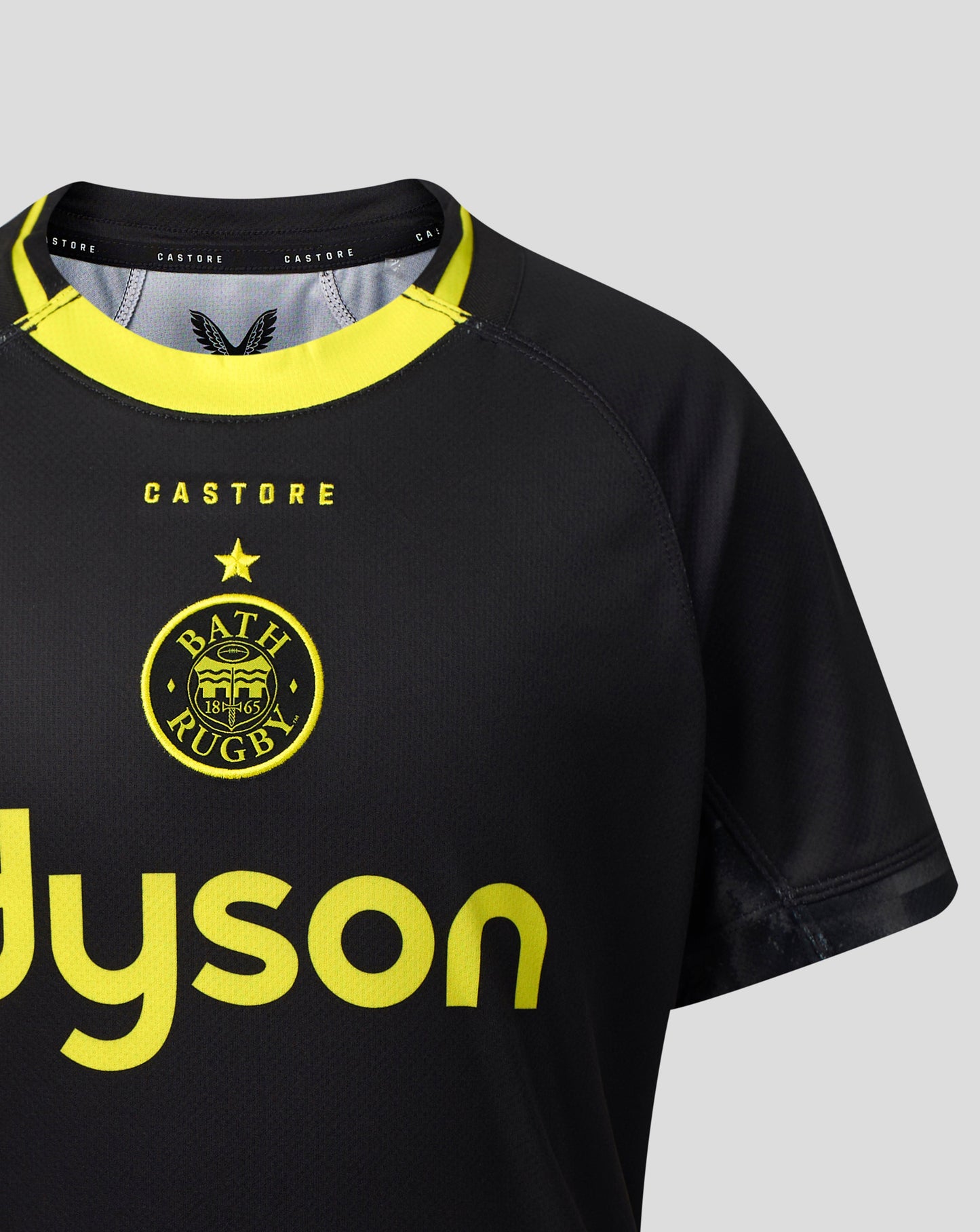 Close-up of a black Bath Rugby jersey with a bright yellow collar, yellow 'CASTORE' text, and a yellow circular Bath Rugby crest on a light gray background.