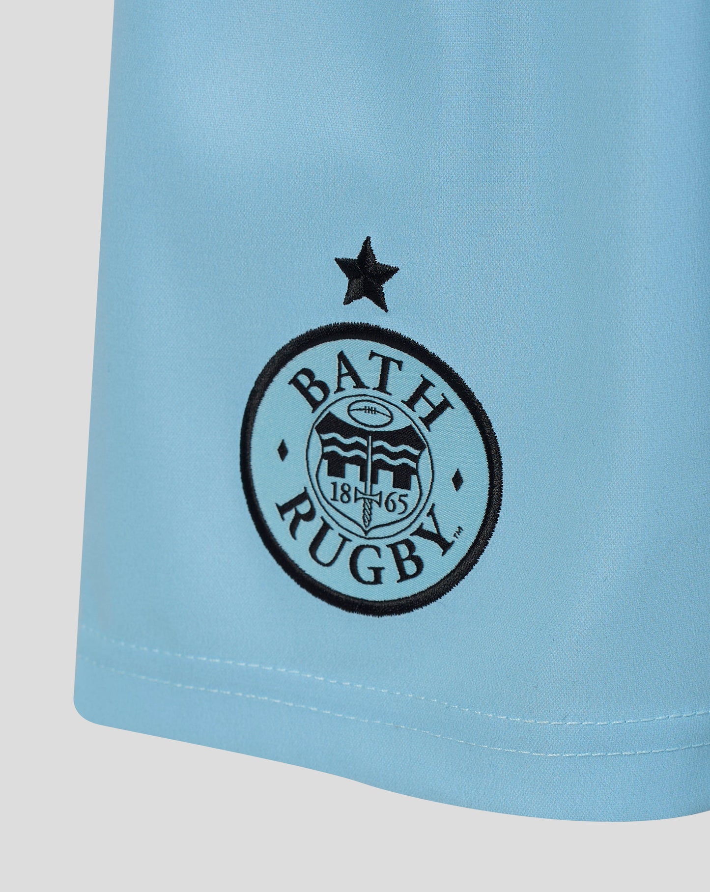 Close-up of a light blue athletic jersey or shorts leg with a black circular Bath Rugby logo and star emblem printed on the front.