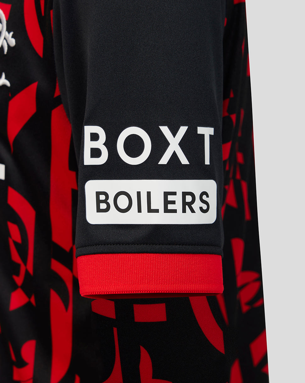 Close-up of a black long-sleeve top with a red abstract pattern, featuring a white sleeve patch that reads 'BOXT BOILERS' and a red ribbed cuff.