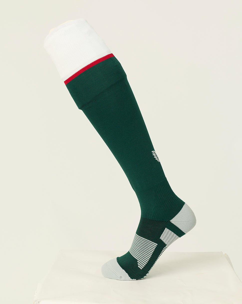 A single knee-high athletic sock displayed on a mannequin leg form against a clean off-white studio background. The sock is dark green with a white cuff, a thin red stripe near the top, and white striped support zones around the heel and toe.