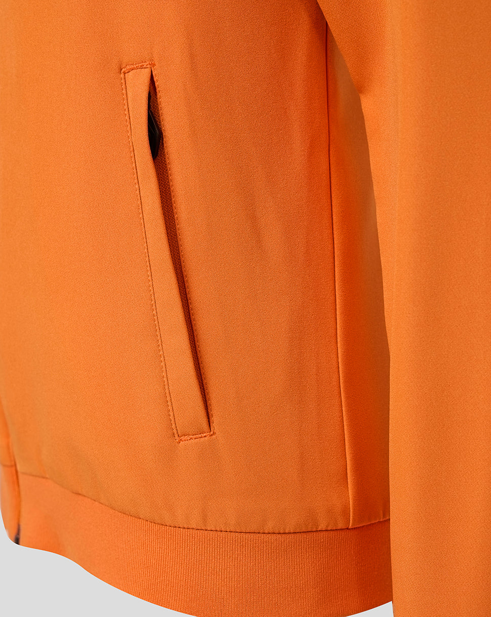 Close-up of an orange jacket or sweatshirt showing a vertical welt pocket and ribbed hem, photographed against a light neutral background.
