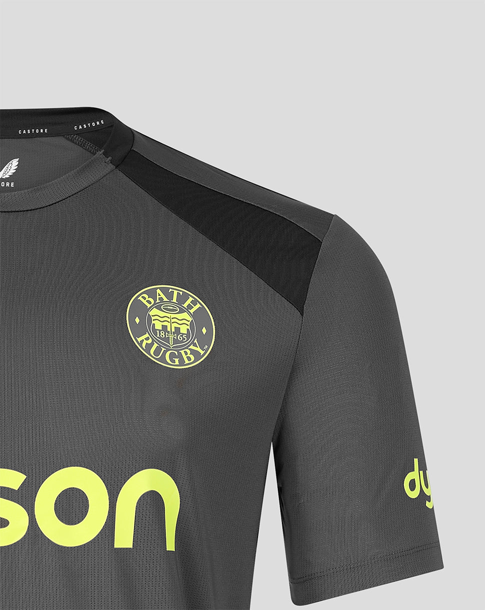 Close-up of a dark gray Bath Rugby athletic jersey with a black shoulder panel, yellow circular club crest, and bright yellow sponsor text on a light gray background.