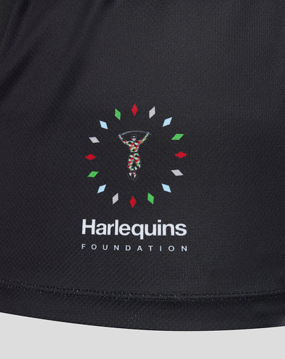 Close-up of a black athletic top featuring the Harlequins Foundation logo printed on the chest. The logo includes a central figure made of colorful dots, surrounded by small multicolored diamond shapes, with 'Harlequins FOUNDATION' printed in white below.