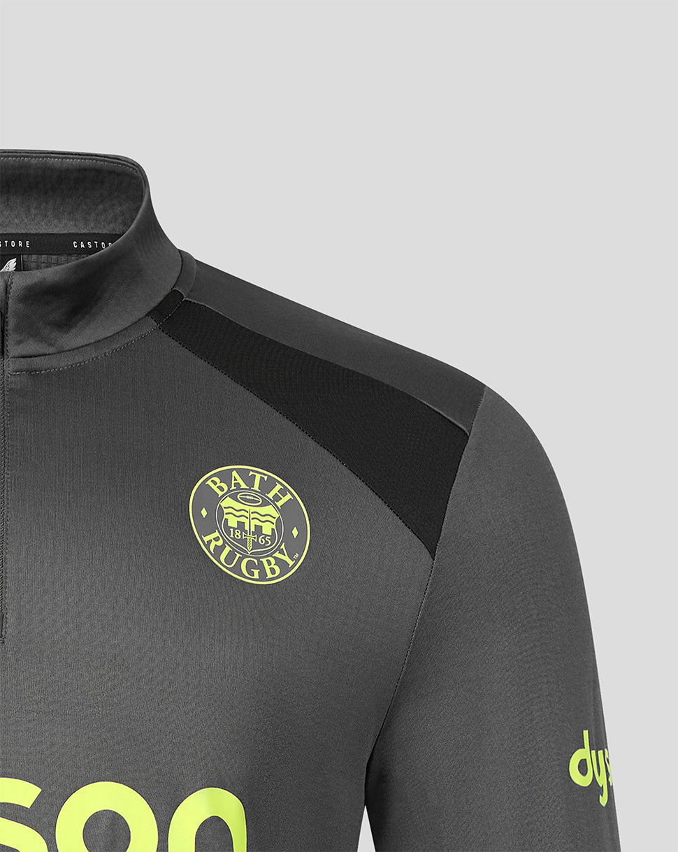 Close-up of a dark gray athletic quarter-zip top with a black shoulder panel, a circular yellow 'Bath Rugby' crest on the chest, and a yellow 'dyne' logo on the sleeve, set against a light gray background.