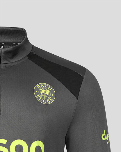 Close-up of a dark gray athletic quarter-zip top with a black shoulder panel, a circular yellow 'Bath Rugby' crest on the chest, and a yellow 'dyne' logo on the sleeve, set against a light gray background.