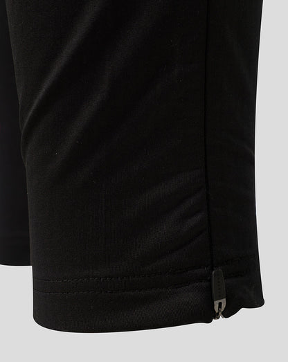 Close-up detail of black athletic joggers showing the hem with a stitched cuff and a small side zipper pull, on a light gray background.