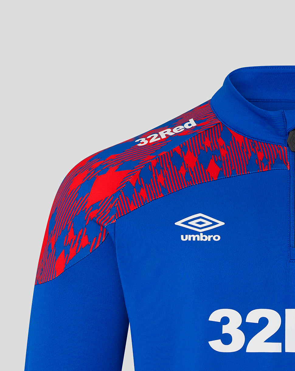 Close-up of a blue and red patterned athletic quarter-zip jacket by Umbro, featuring the white Umbro logo and '32Red' text on the shoulder, set against a light gray background.