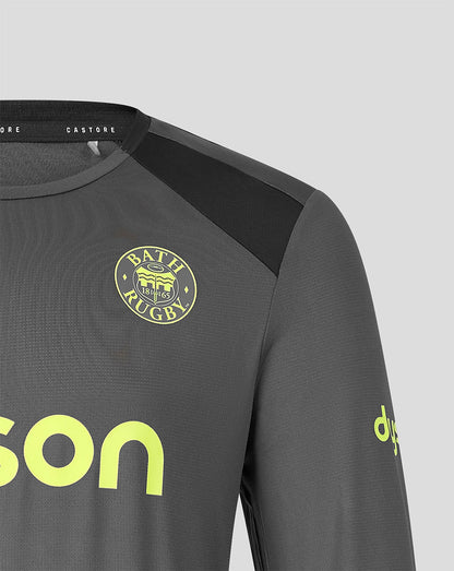 Close-up of a dark gray long-sleeve sports jersey with a black shoulder panel, a circular Bath Rugby crest on the chest, and neon yellow-green branding details on a light gray background.