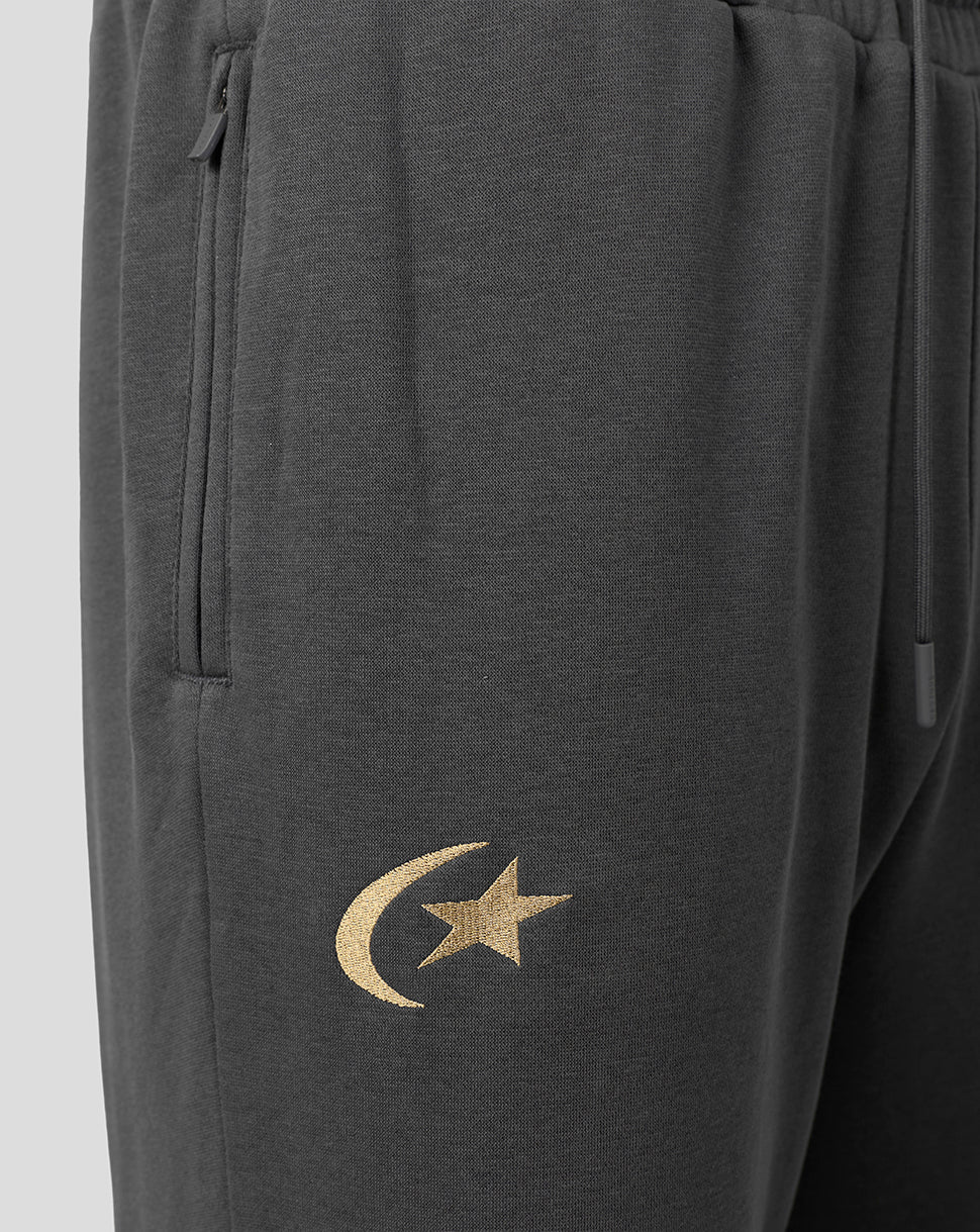 Close-up of dark gray sweatpants featuring a gold embroidered crescent moon and star logo on the lower left leg, with a side zip pocket and drawstring waistband.