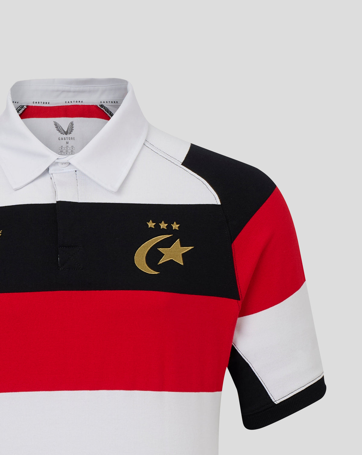 A close-up of a short-sleeve rugby polo shirt with horizontal black, red, and white color-blocked stripes and a gold crescent and star emblem on the chest, shown on a light gray background.