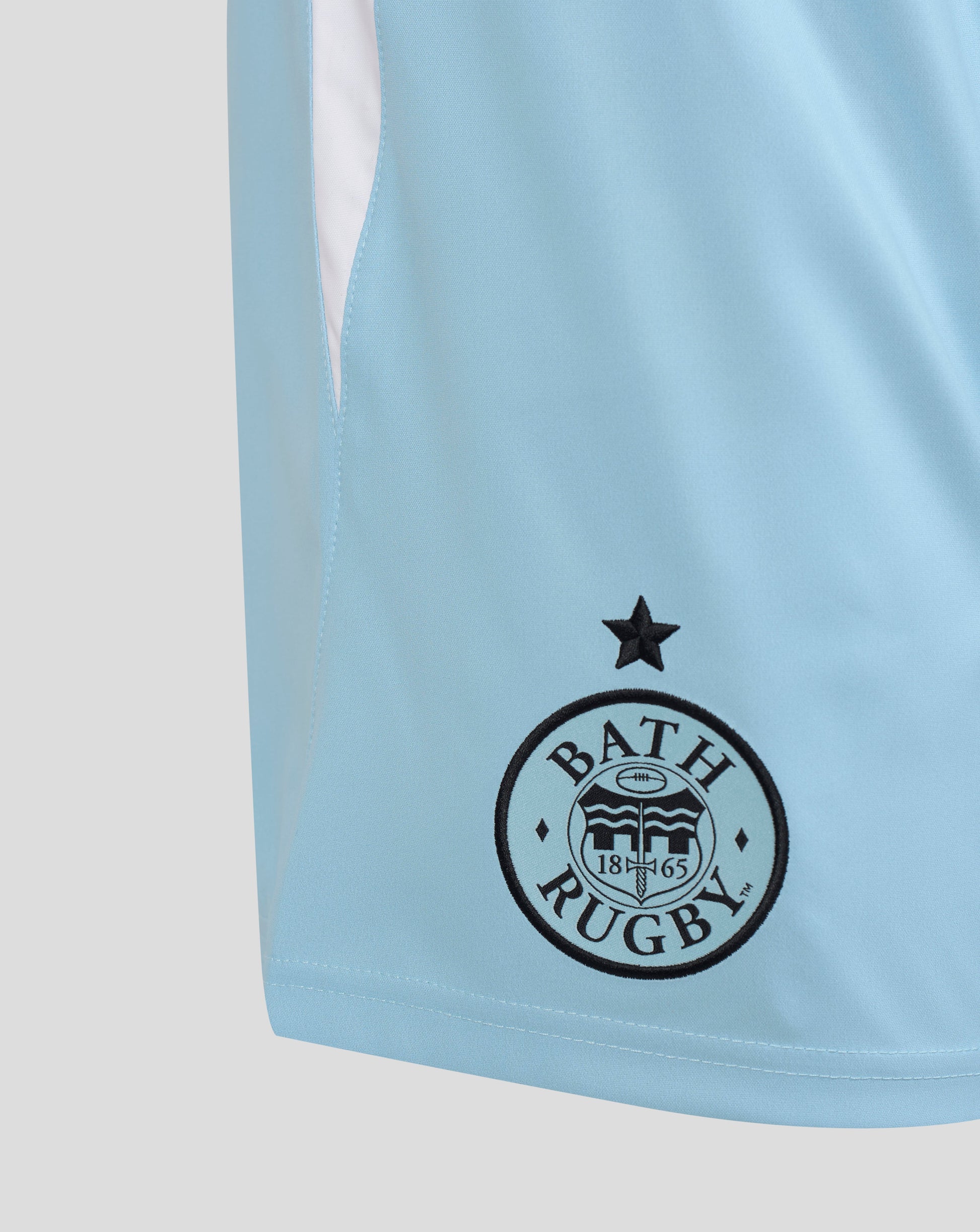Close-up of a light blue athletic jersey showing the black Bath Rugby club crest on the front, with a white side panel visible at the upper left.