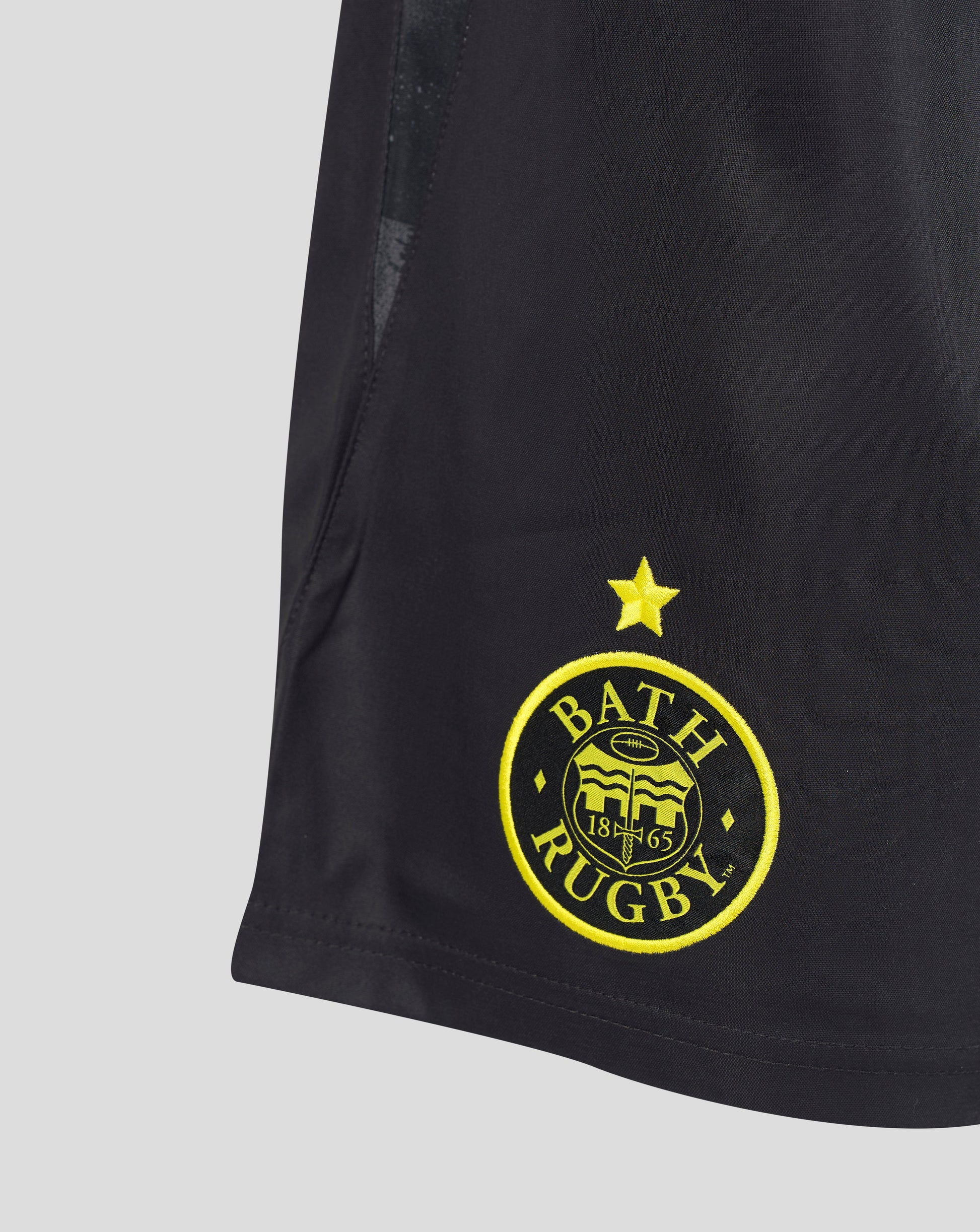 Close-up of a black athletic shorts leg featuring a yellow embroidered Bath Rugby club crest and star on the lower left side, set against a light gray background.