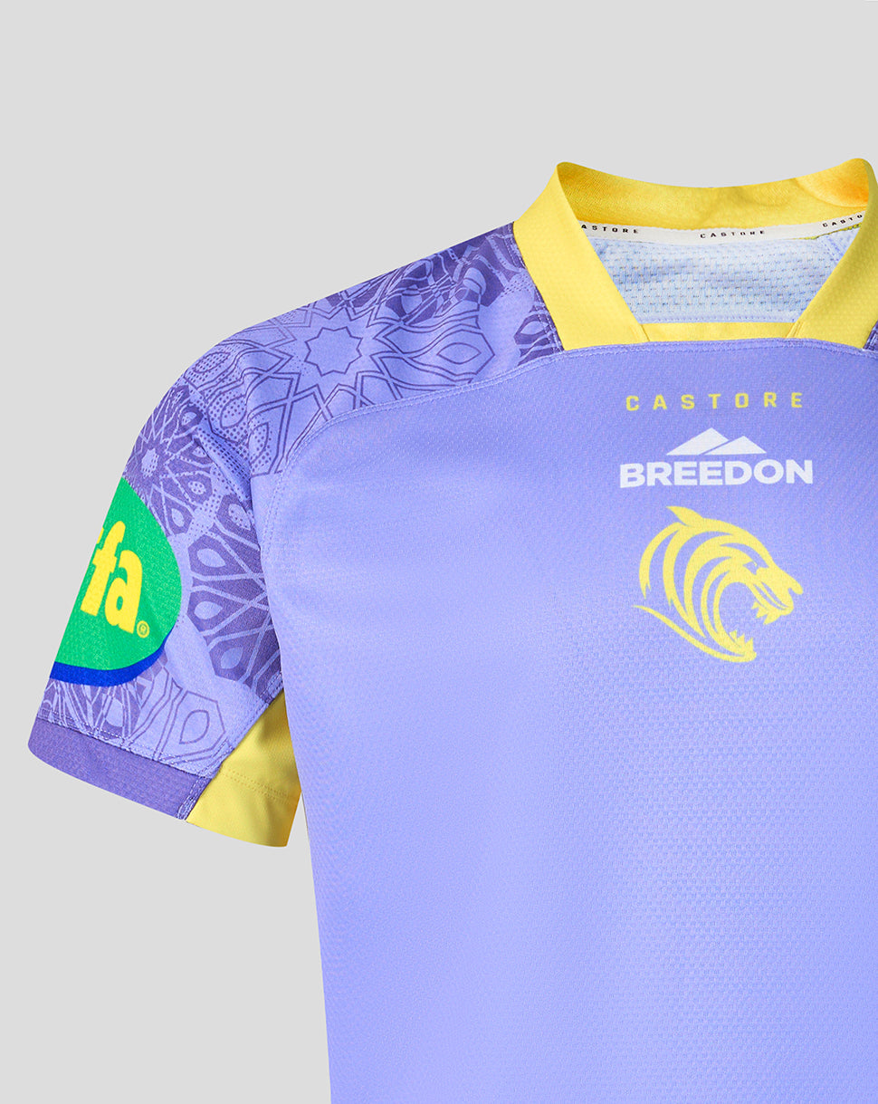 A close-up of a short-sleeve rugby jersey in light purple with yellow trim, featuring a purple patterned right sleeve, a green oval sponsor patch on the sleeve, and yellow and white printed branding on the chest.