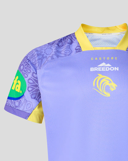 A close-up of a short-sleeve rugby jersey in light purple with yellow trim, featuring a purple patterned right sleeve, a green oval sponsor patch on the sleeve, and yellow and white printed branding on the chest.