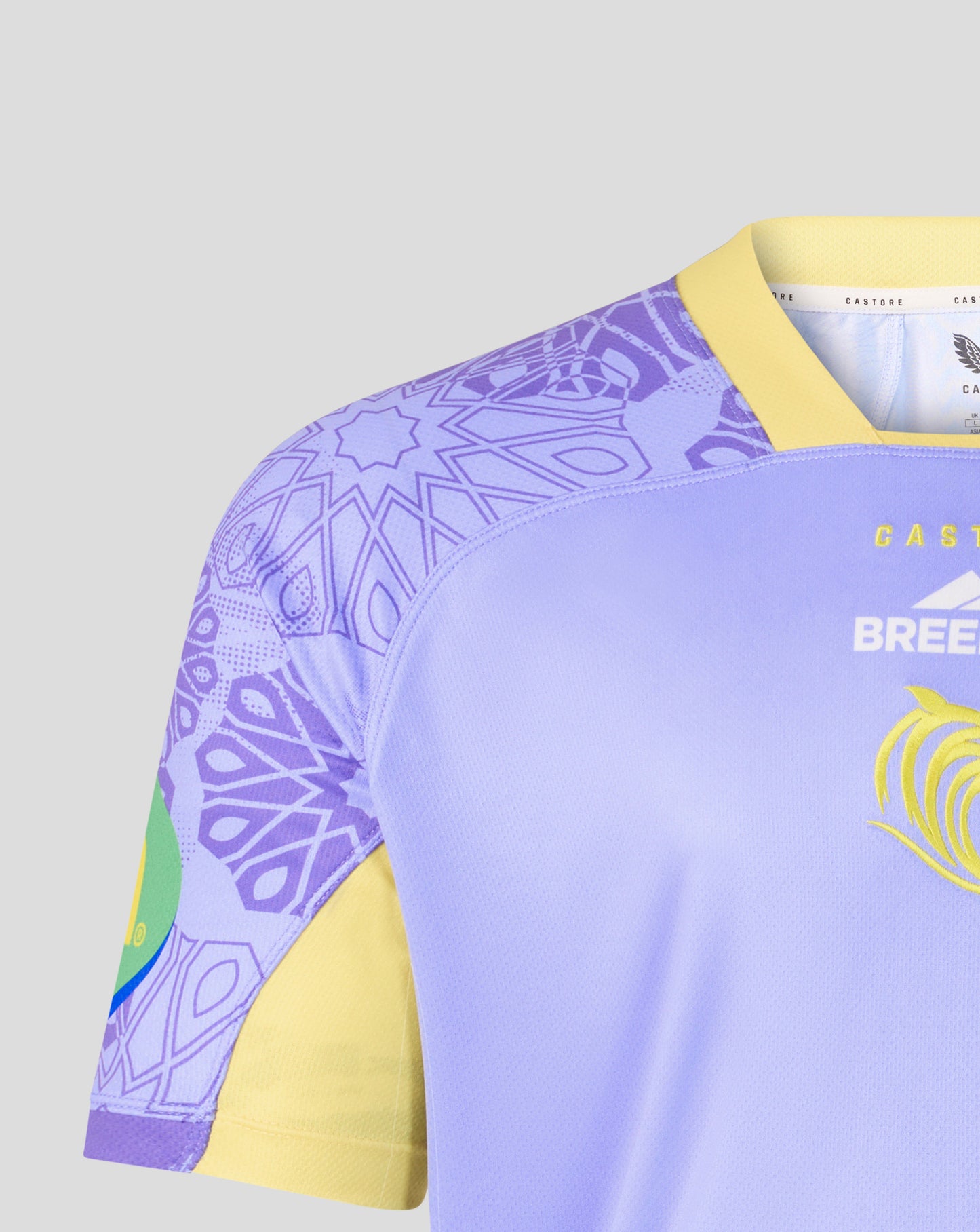 Close-up of a rugby jersey showing the shoulder and upper chest area. The jersey has a light purple body, a yellow V-neck collar and sleeve cuffs, a purple geometric pattern on the shoulder, and a yellow graphic on the chest.