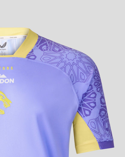 Close-up of a soccer jersey showing the shoulder and sleeve area. The jersey is light purple with a yellow collar and sleeve cuff, featuring a purple geometric pattern on the upper sleeve and shoulder, and yellow accents on the side.
