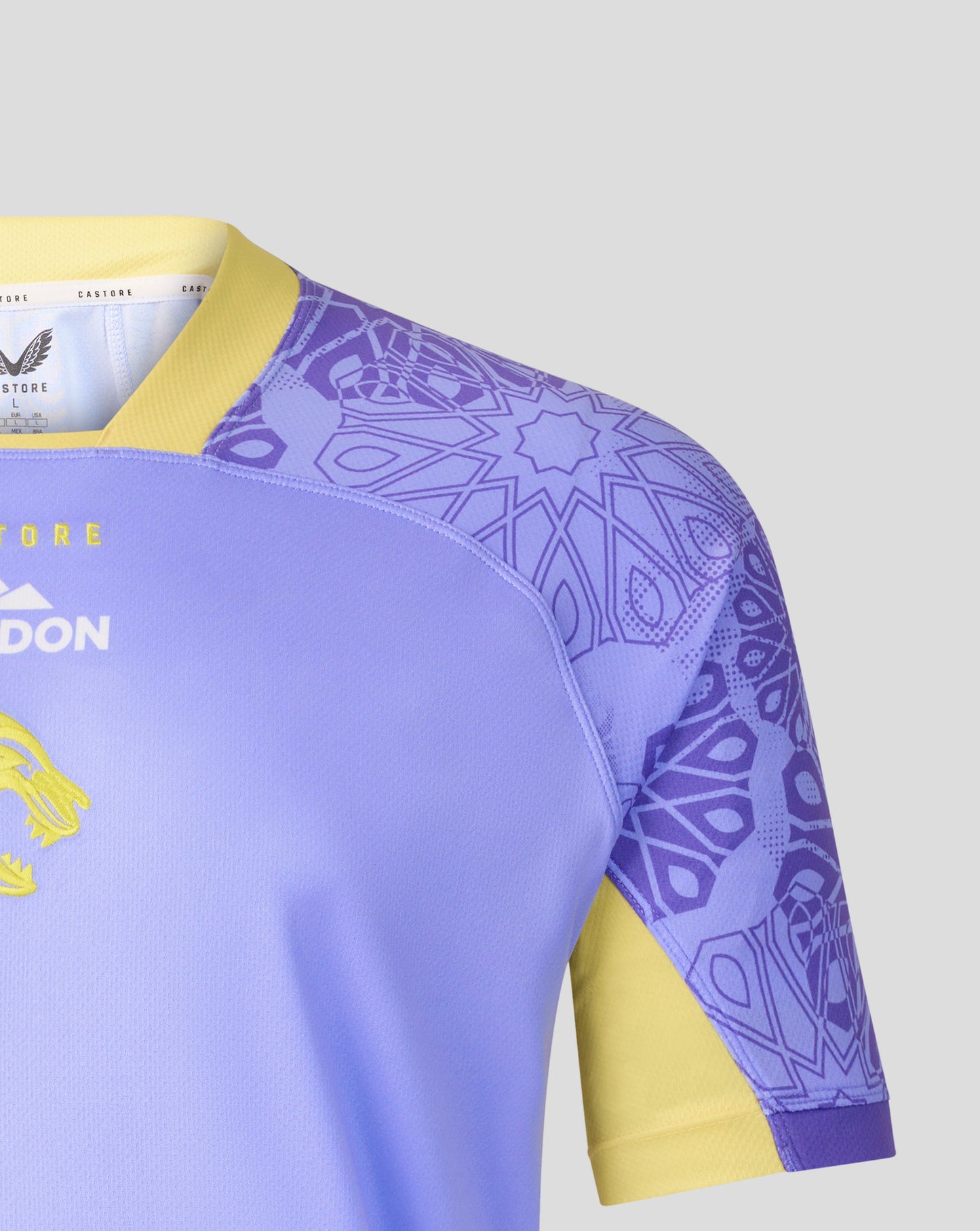 Close-up of a rugby jersey showing the shoulder and sleeve area. The jersey is light purple with a yellow collar and sleeve cuff, featuring a purple geometric pattern on the upper sleeve and shoulder, and yellow accents on the side.
