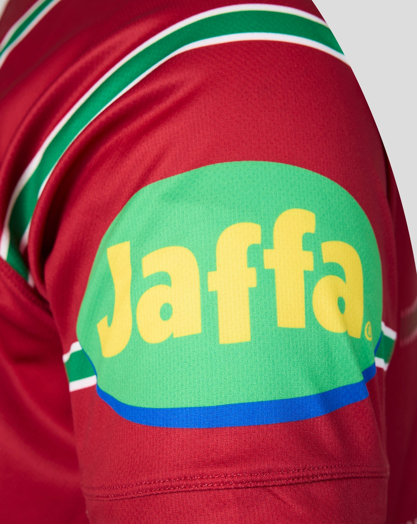 Close-up of a red sports jersey sleeve featuring a large green oval patch with yellow 'Jaffa' lettering and green, white, and blue stripe details.