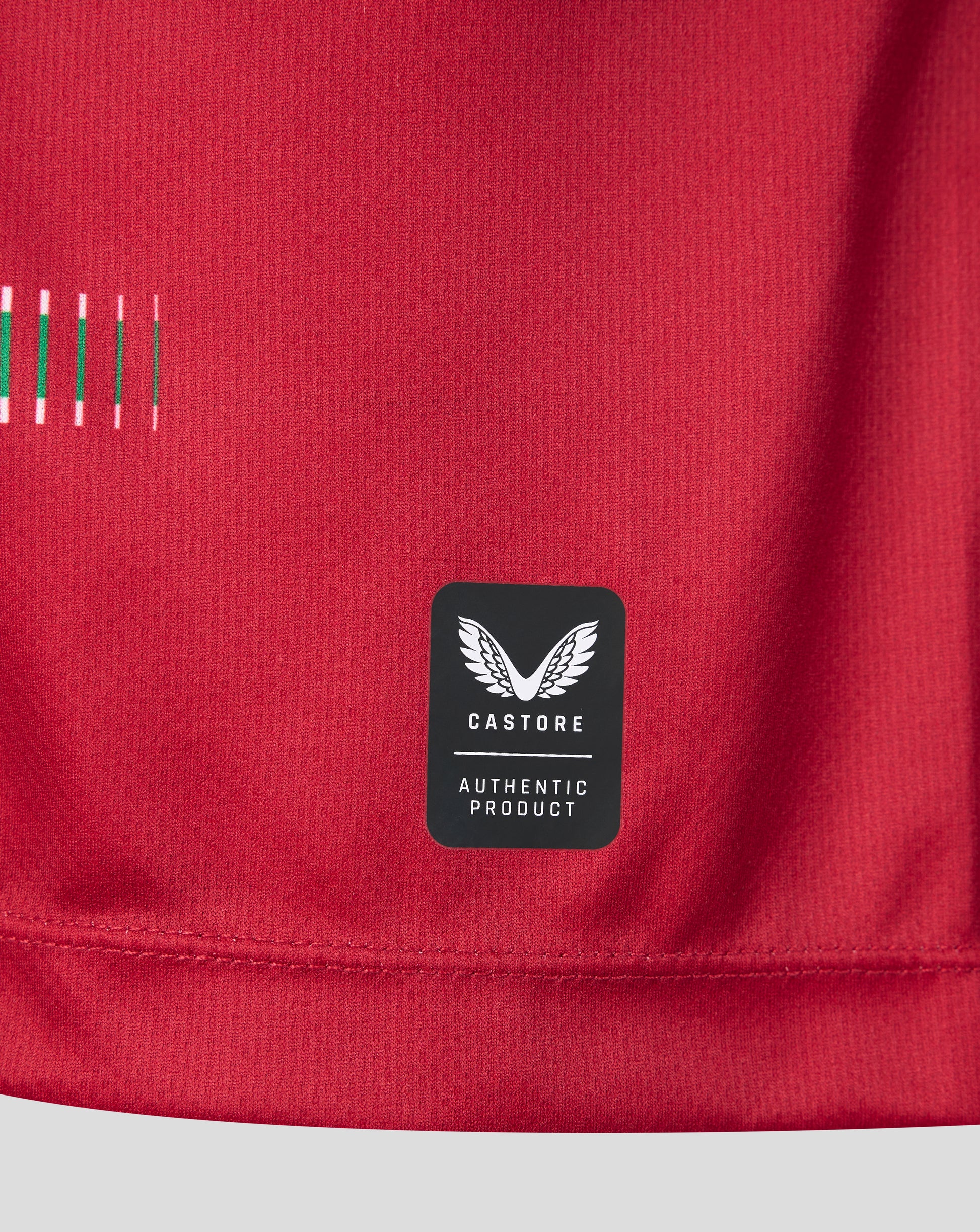 Close-up of a red athletic top showing a black Castore brand patch with white wing logo and 'AUTHENTIC PRODUCT' text, plus a small striped detail on the left side.