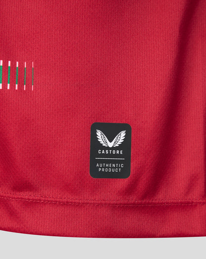 Close-up of a red athletic top showing a black Castore brand patch with white wing logo and 'AUTHENTIC PRODUCT' text, plus a small striped detail on the left side.