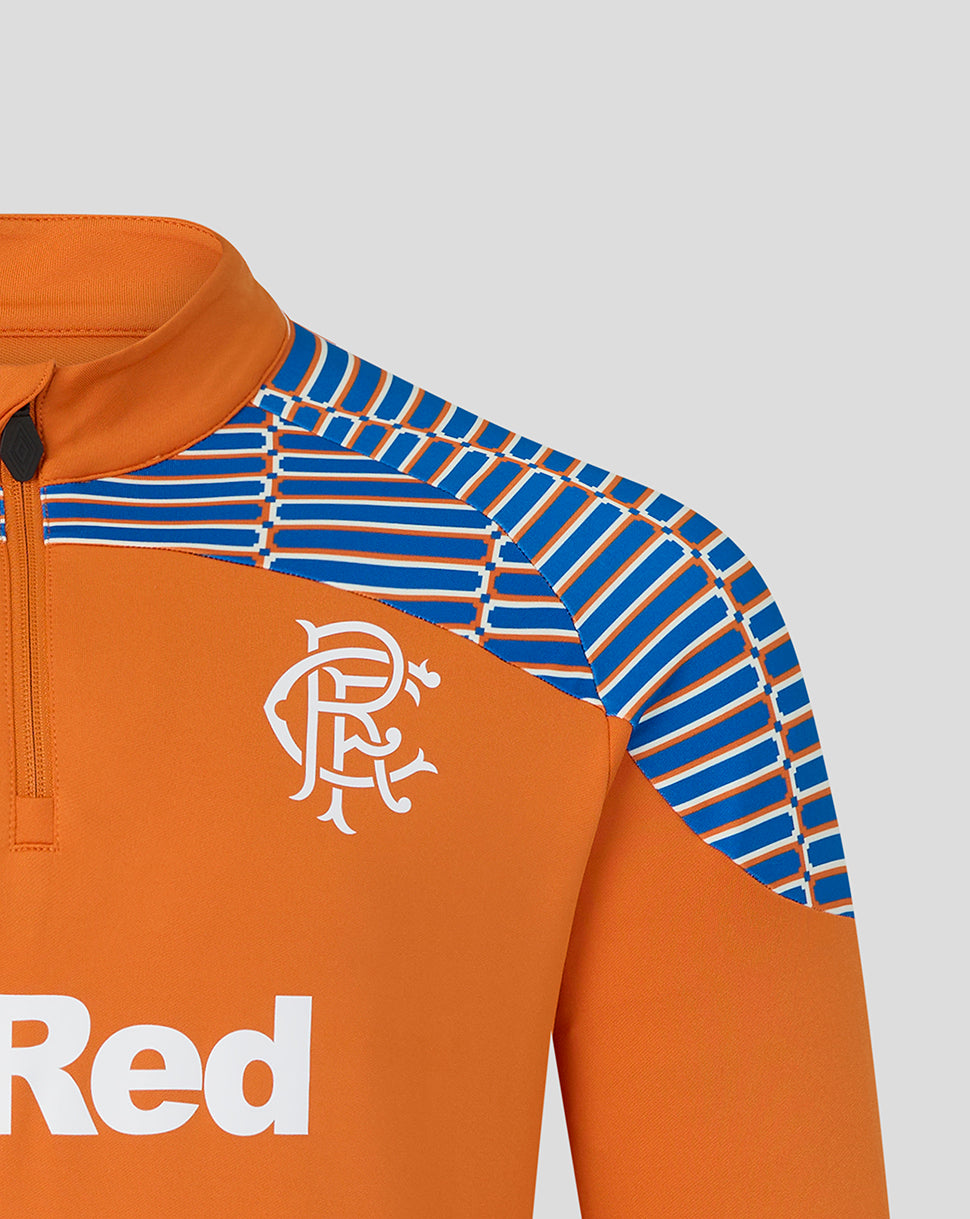 Close-up of an orange athletic quarter-zip top with a blue and white striped patterned shoulder panel and a white stylized crest on the chest, shown against a light gray background.