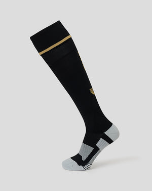 A single knee-high athletic sock shown on a light gray background. The sock is primarily black with a thin gold stripe near the cuff, a small gold logo on the side, and gray striped detailing on the toe and heel.