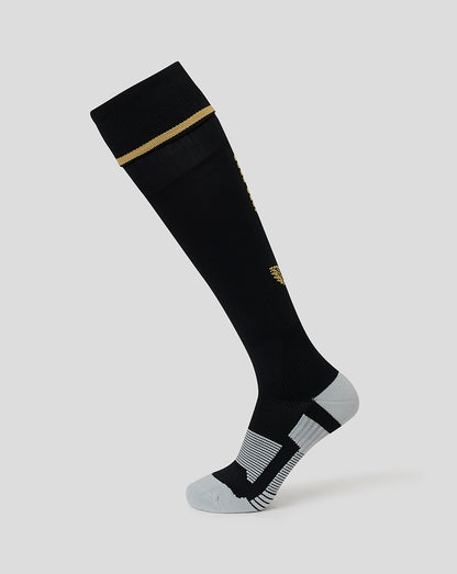 A single knee-high athletic sock shown on a light gray background. The sock is primarily black with a thin gold stripe near the cuff, a small gold logo on the side, and gray striped detailing on the toe and heel.