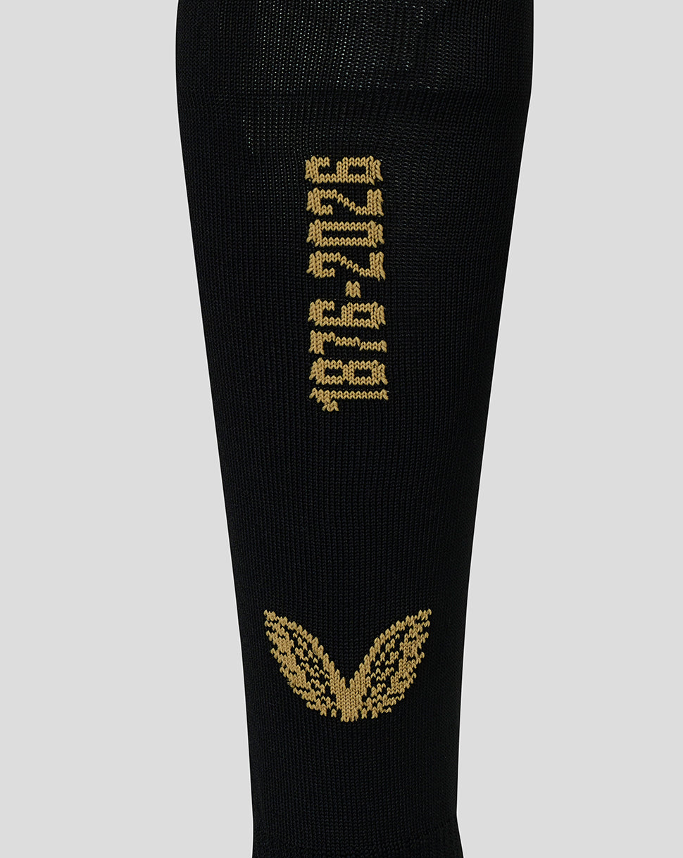 A close-up of a black knit sock featuring gold knitted text reading '1976-2026' and a gold winged emblem near the bottom, shown against a light gray background.