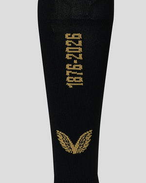 A close-up of a black knit sock featuring gold knitted text reading '1976-2026' and a gold winged emblem near the bottom, shown against a light gray background.