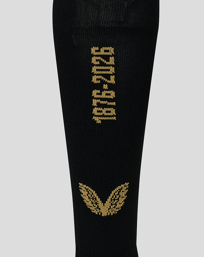 A close-up of a black knit sock featuring gold knitted text reading '1976-2026' and a gold winged emblem near the bottom, shown against a light gray background.