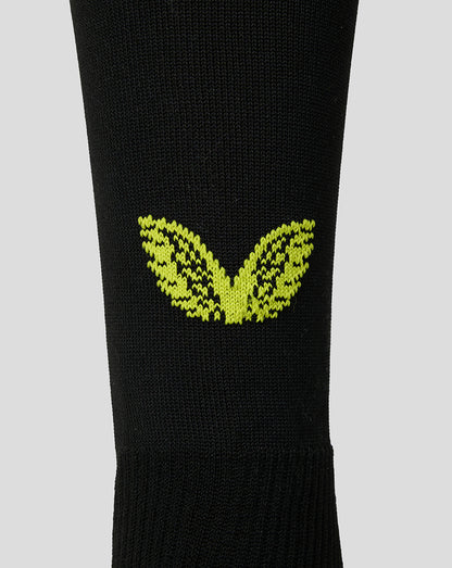 A close-up of a black knit sock featuring a bright yellow-green embroidered winged logo on the calf, set against a solid light gray background.