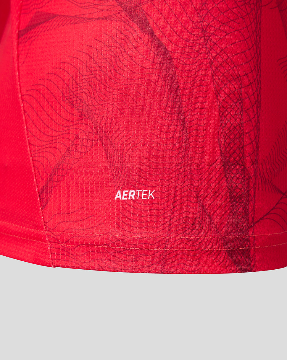 Close-up of a red athletic garment showing the AERTEK logo printed in white on the fabric, with a subtle dark wavy pattern over a bright red mesh-like surface.