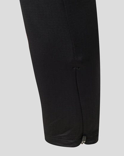 Close-up of the lower leg of black pants showing a side seam and a small metal zipper pull at the hem, set against a solid light gray background.