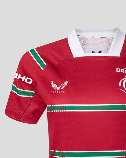 A close-up of a red Castore rugby jersey with green and white stripes, featuring the Castore wing logo on the chest and a sponsor logo on the sleeve, set against a solid gray background.