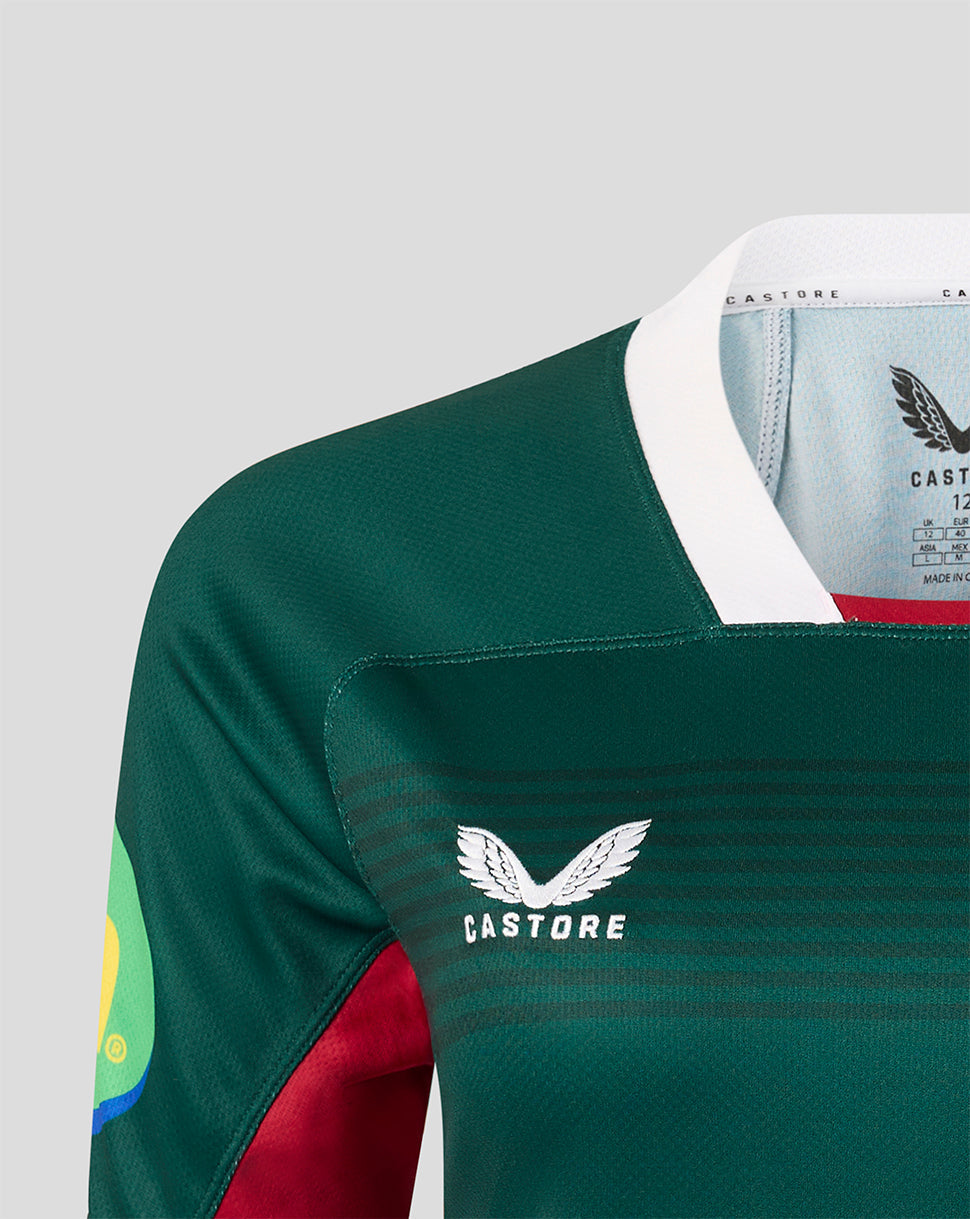 Close-up of a Castore rugby jersey in dark green with a white collar, white chest logo, and red sleeve panel, shown on a light gray background.