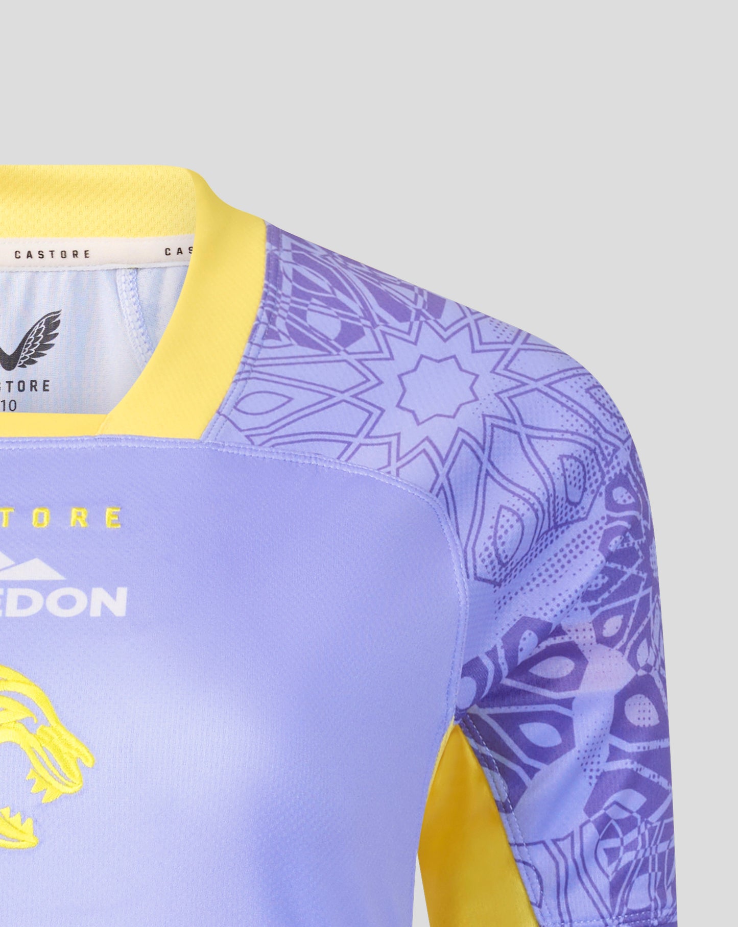 Close-up of a soccer jersey with a light purple body, yellow collar and sleeve accents, and a purple geometric pattern on the sleeves. The brand Castore is visible on the inner neck label.