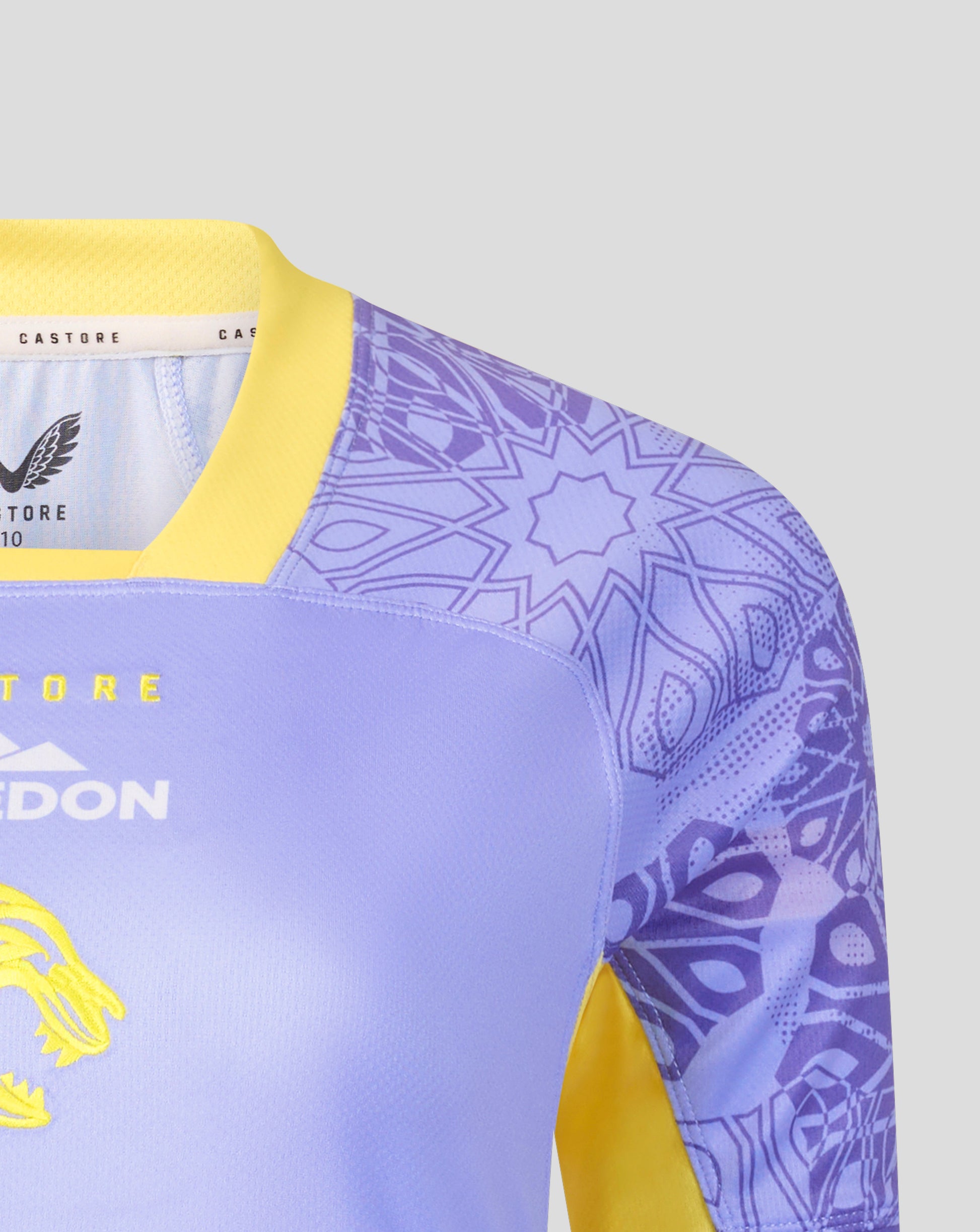 Close-up of a soccer jersey with a light purple body, yellow collar and sleeve accents, and a purple geometric pattern on the sleeves. The brand Castore is visible on the inner neck label.