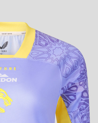 Close-up of a soccer jersey with a light purple body, yellow collar and sleeve accents, and a purple geometric pattern on the sleeves. The brand Castore is visible on the inner neck label.